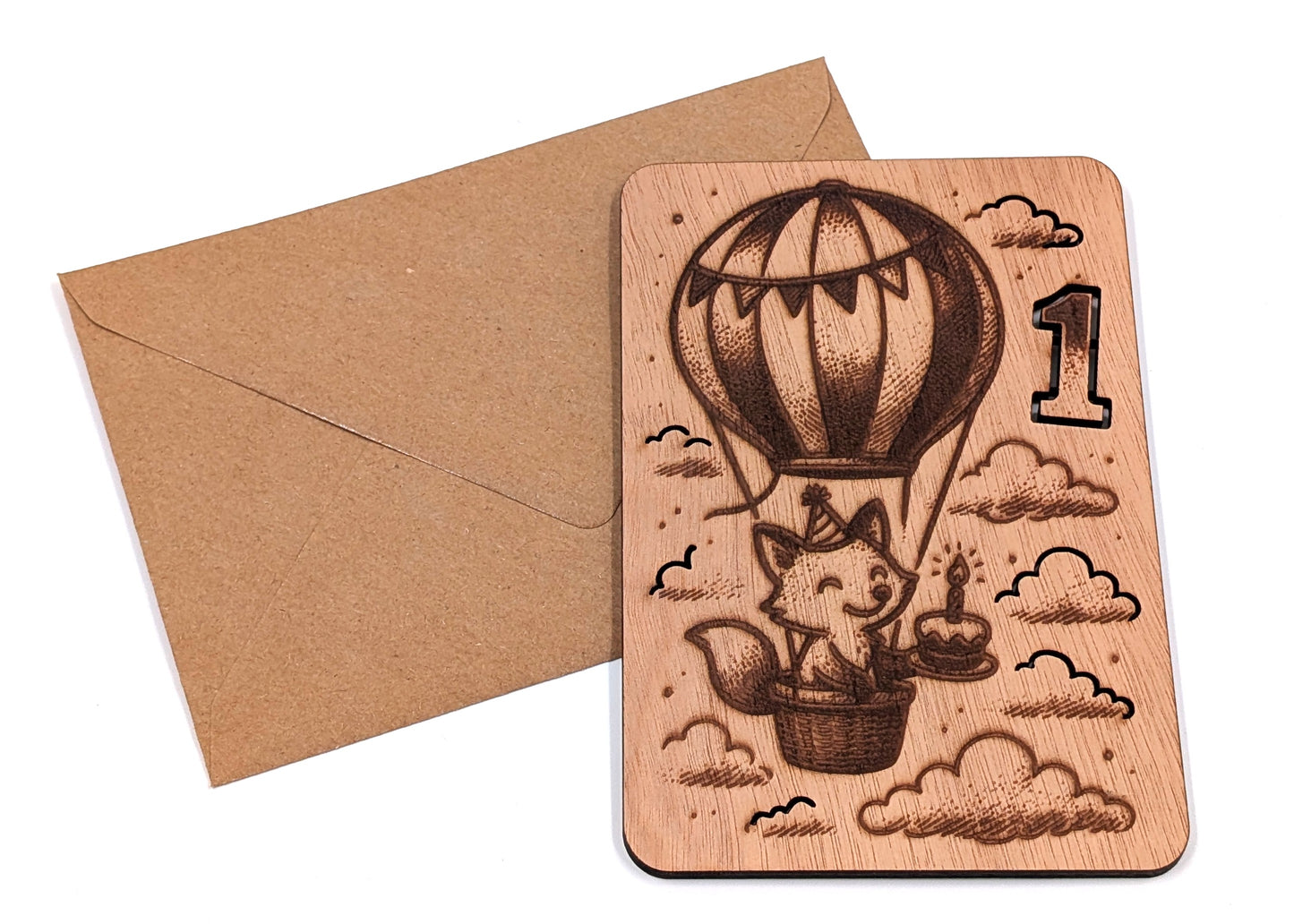 Handgemaakte houten kaart voor een eerste verjaardag met luchtballon-thema naast een bruine kraft envelop op een witte achtergrond.