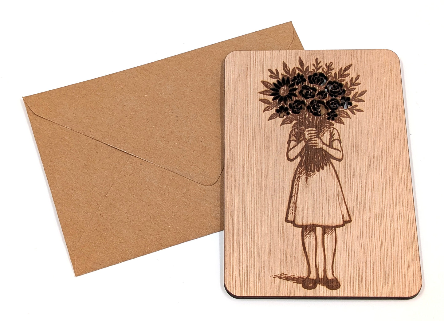 Handgemaakte houten kaart met een artistiek bloemenmeisje-ontwerp naast een duurzame bruine kraft envelop op een witte achtergrond.