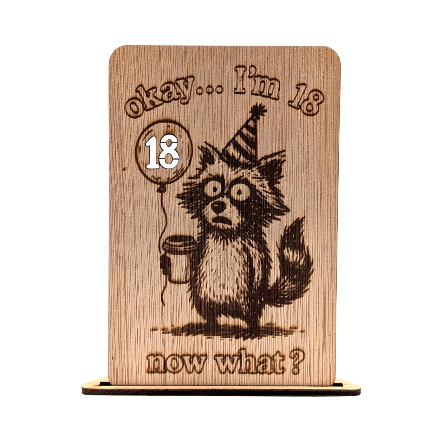 Houten verjaardagskaart 18 jaar met een grappige gravure van een wasbeer met feestmuts en koffie, inclusief de tekst 'okay... I'm 18 now what?'.